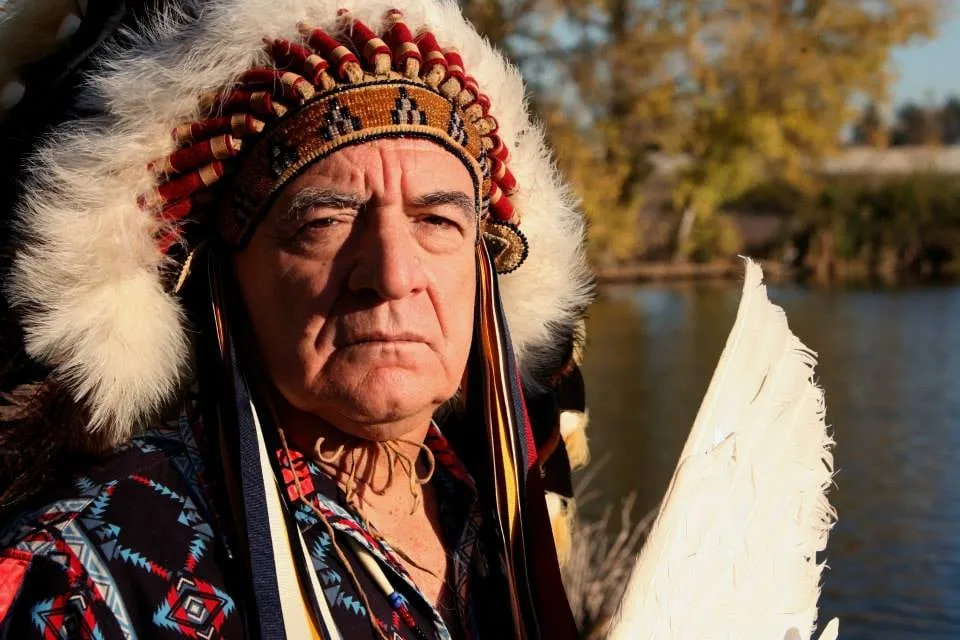 An elder in a feathered headdress with red and white beadwork and a patterned shirt holds a white feather fan beside a lake with autumn trees.