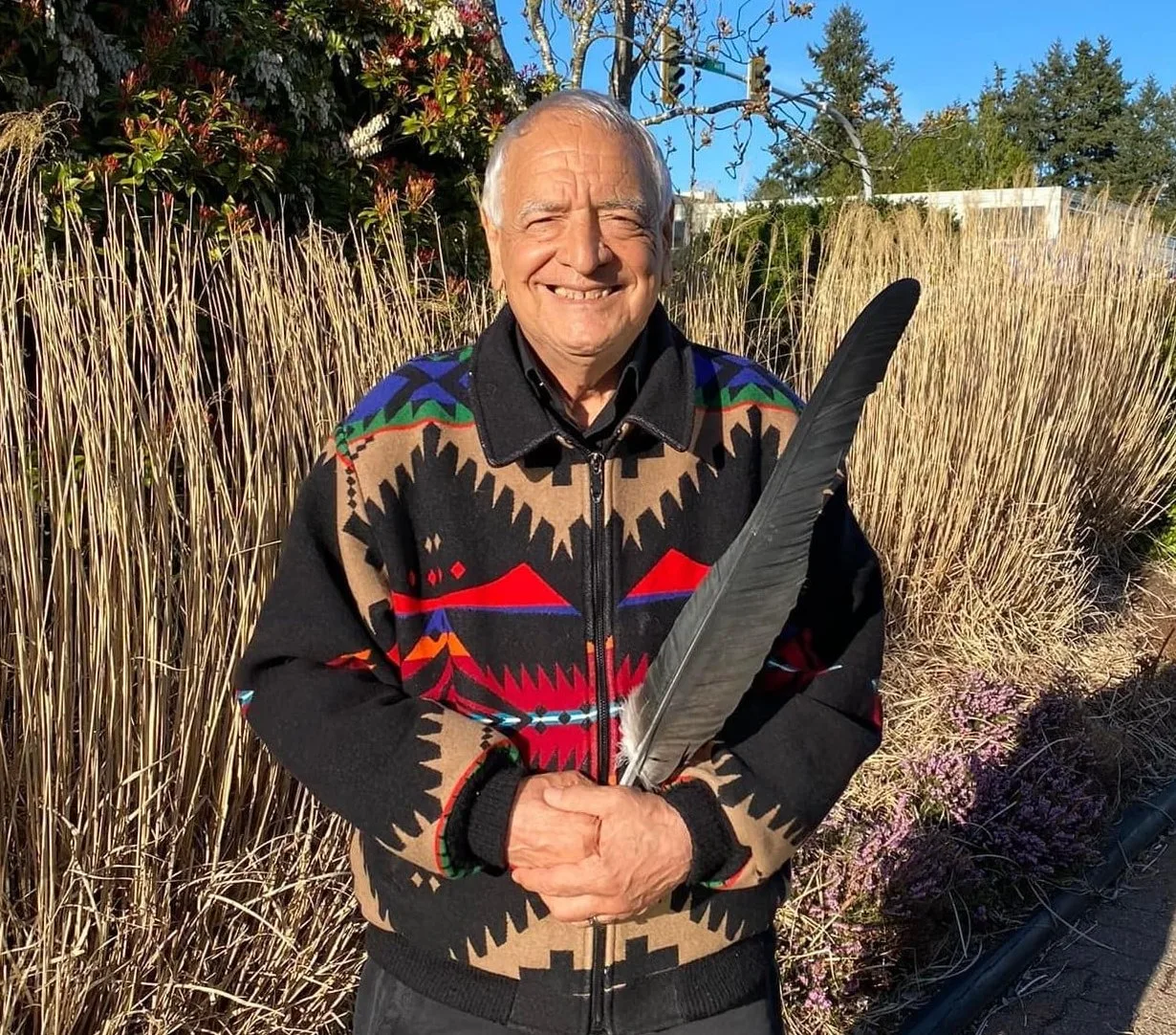 A smiling elder with white hair, wearing a black jacket with colorful geometric patterns, holds a large dark condor feather in front of tall dry grasses on a sunny day.
