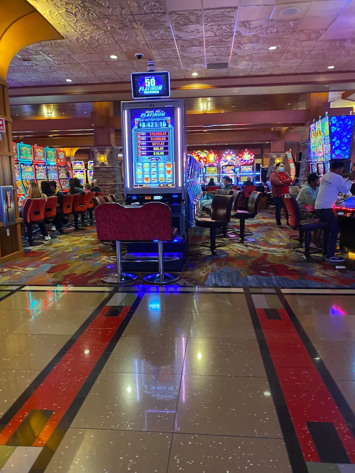 A wide casino floor filled with glowing slot machines and patrons, a tall Quick Hit Platinum machine showing jackpot amounts in the center.