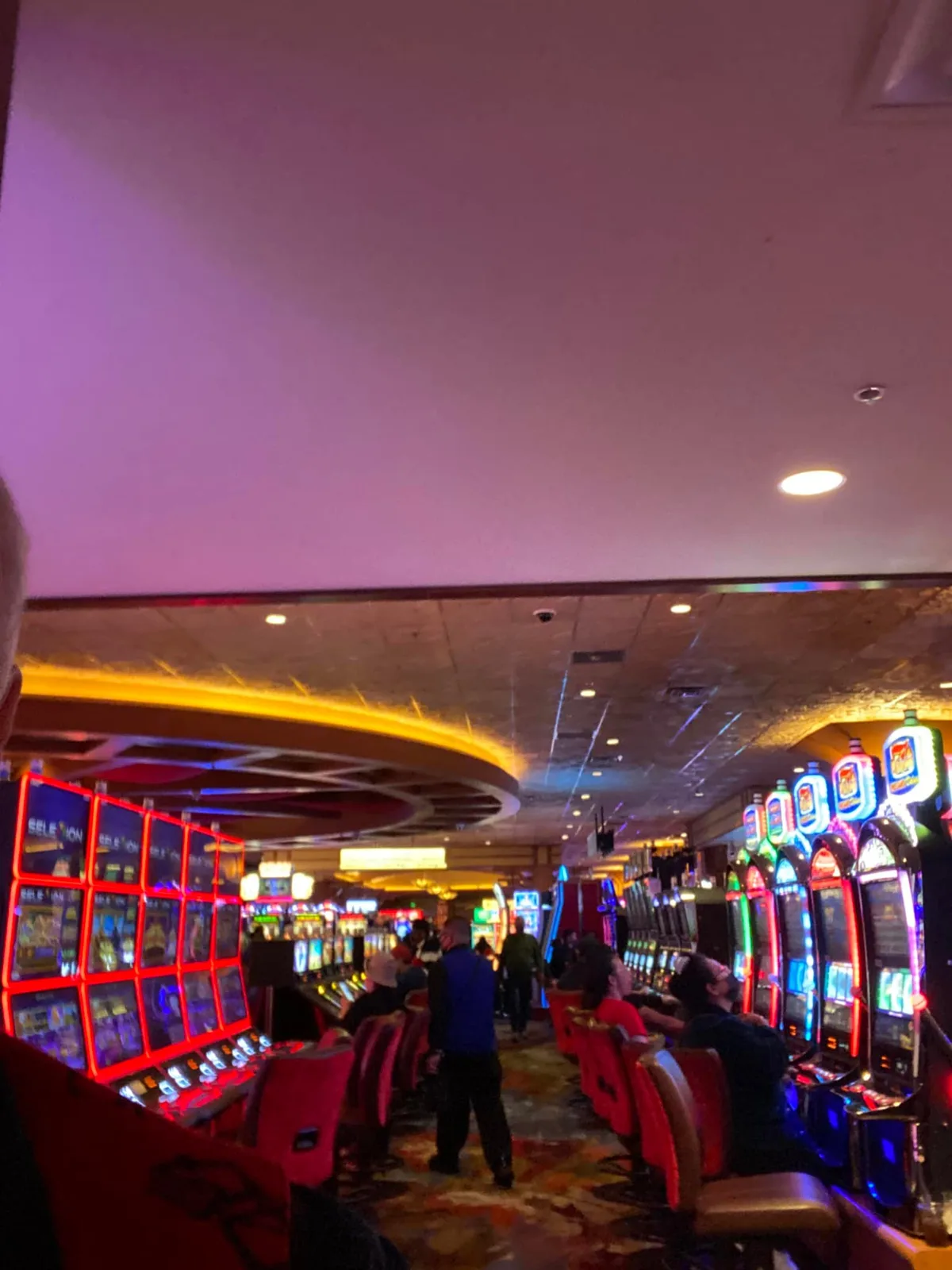 A long aisle of flashing slot machines with people seated at them and others walking, under a purple-lit ceiling and an amber-lit overhead ring.