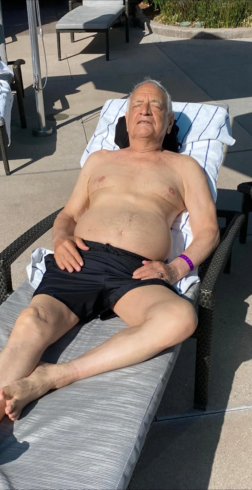 An elderly man with white hair, shirtless in black swim shorts, rests with eyes closed on a poolside lounge chair in bright sunshine, wearing a purple wristband.