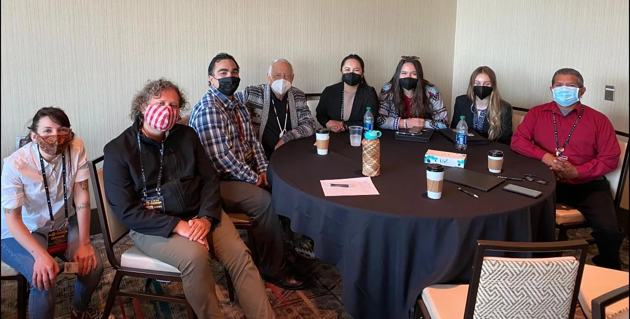 Eight people wearing face masks and conference lanyards sit around a round black-draped table with coffee cups and papers in a meeting room, including an elder in a patterned sweater.
