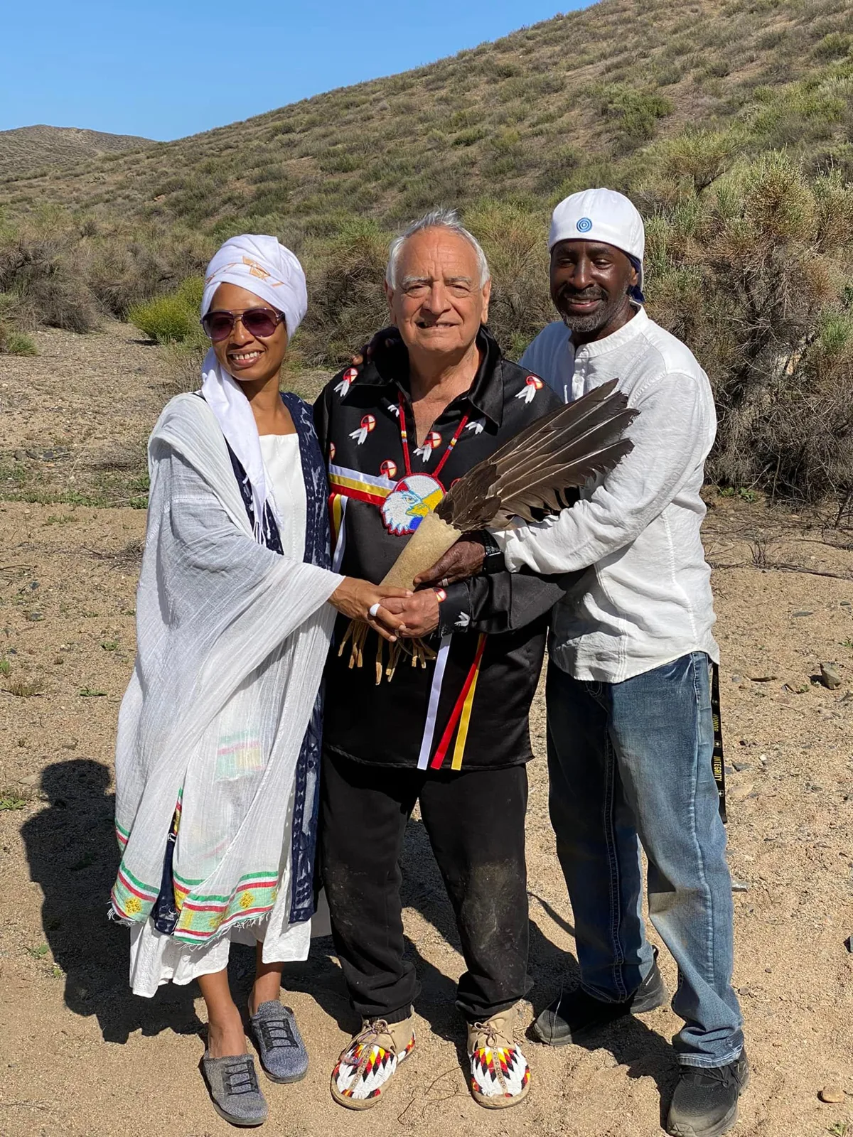 Phil Lane Jr., in a black ribbon shirt with a beaded eagle medallion, holds an eagle feather fan with Sister Antoinette and Brother Pato, both in white, on a desert hillside.