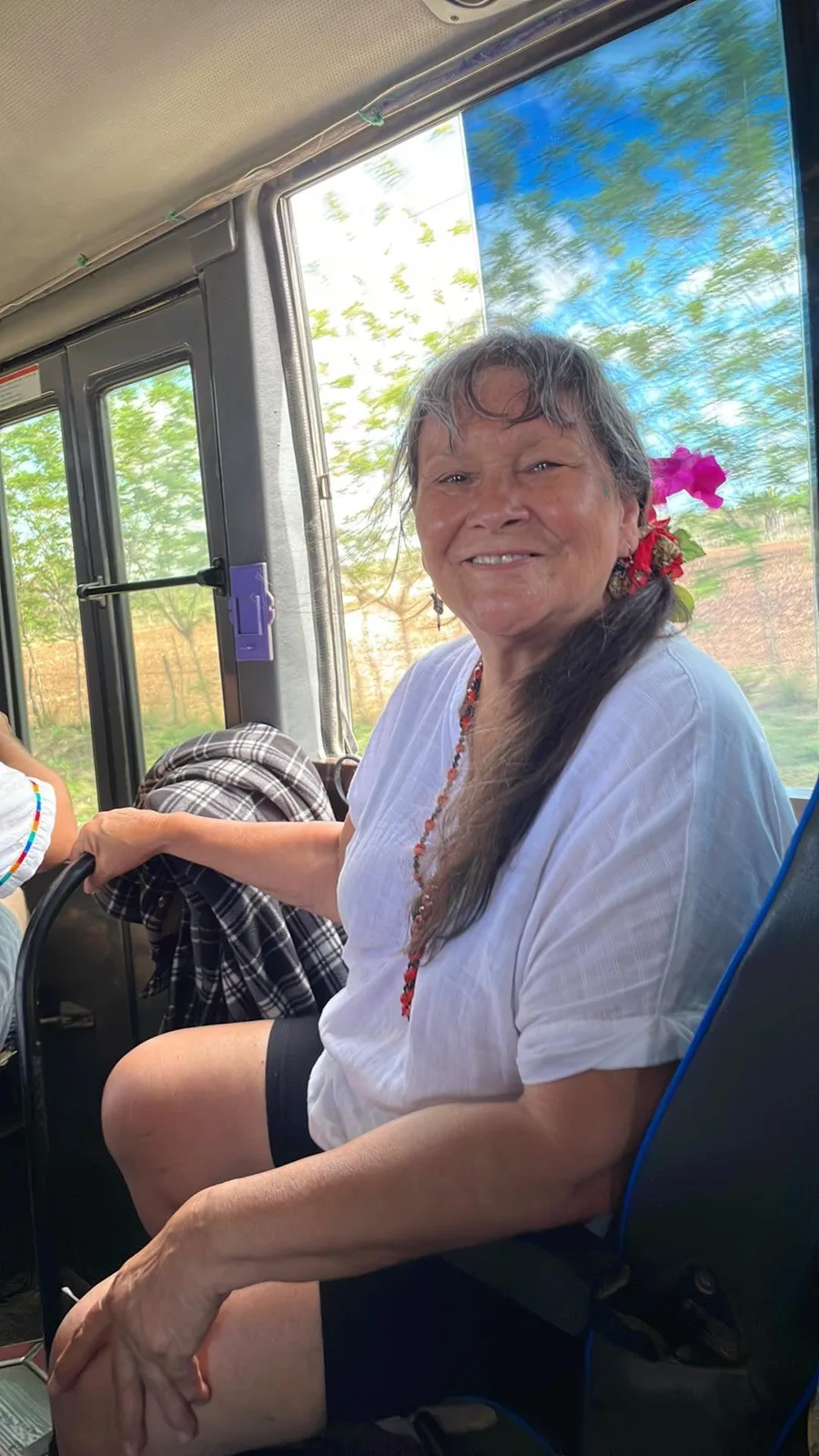 A smiling woman with long gray-streaked hair and a pink flower tucked behind her ear sits on a bus in a white blouse and red beaded necklace, green trees blurring past the window.