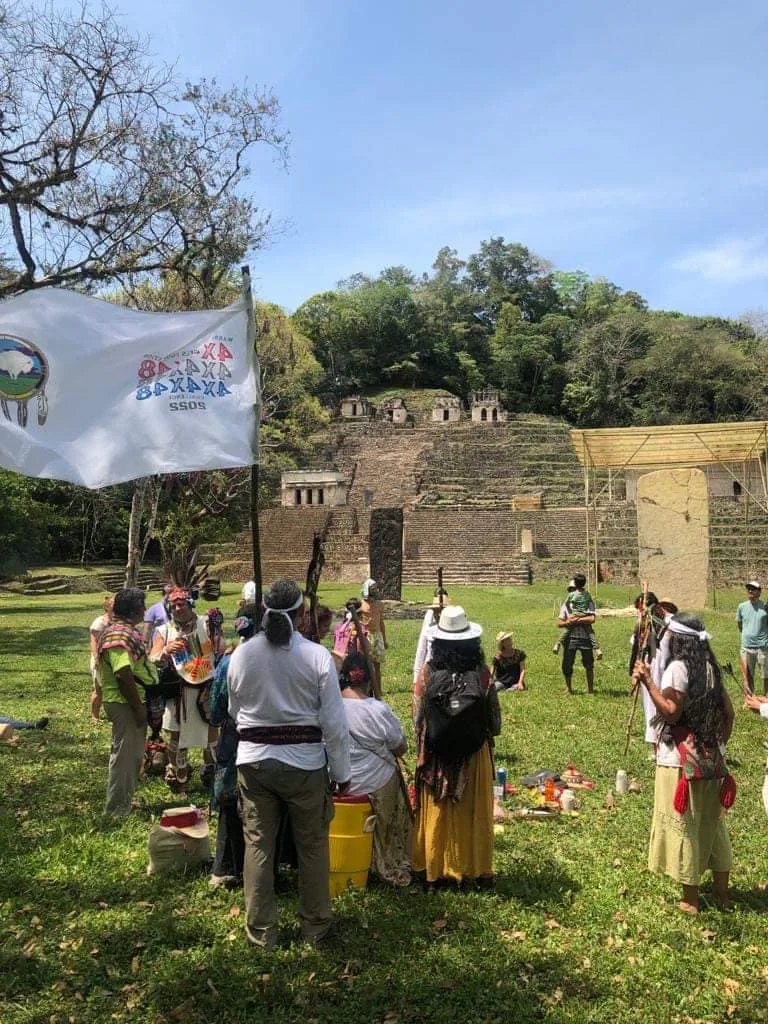 A circle of people in traditional dress gathers on a grassy field before terraced Maya ruins, with a white flag showing a white buffalo and 4x4x48 lettering in the foreground.