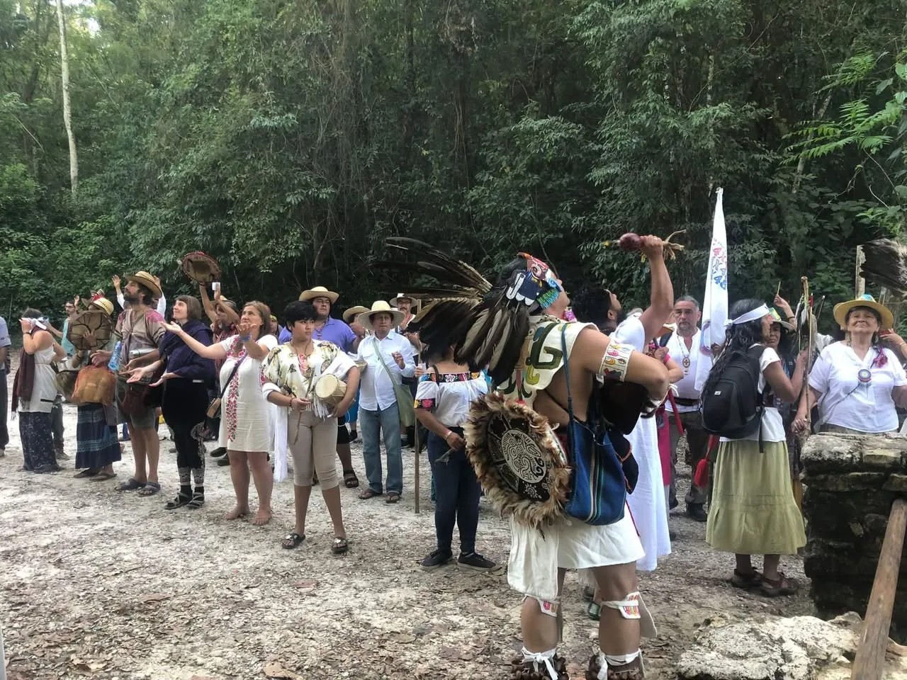 A large group in white and embroidered clothing raises hands skyward in a forest clearing, led by a dancer in a feathered headdress holding up a rattle.