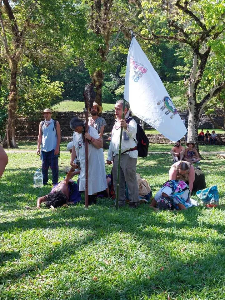 A woman holding a tall eagle staff and a man holding a white 4x4x48 flag with a white buffalo emblem stand on grass while others kneel and bow to the ground.