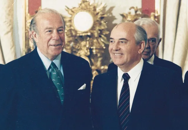 George P. Shultz and Mikhail Gorbachev, both in dark suits, smiling side by side in an ornate room with gilded decor, another man behind them.