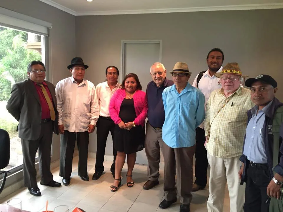 The same group posed in a room with a large window and greenery outside; a woman in a pink cardigan stands near the center among men in shirts and hats.