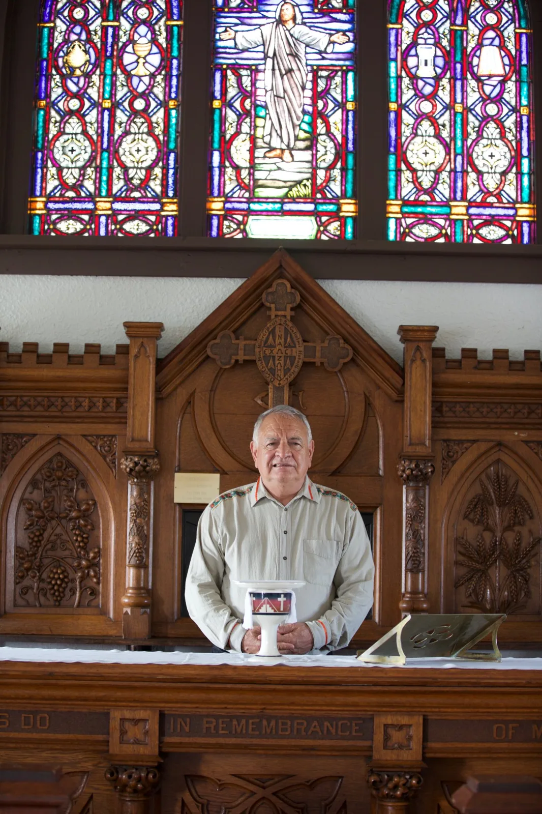 Phil Lane Jr., a white-haired man in a light shirt with beaded trim, stands behind a carved wooden church altar holding a chalice, beneath stained glass windows depicting Jesus.