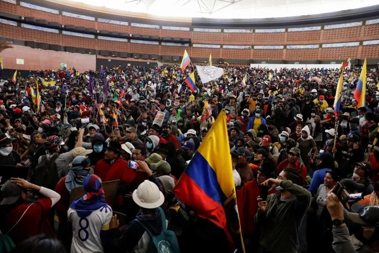 A huge crowd of Indigenous protesters fills an indoor arena, many wearing red ponchos and hats, waving Ecuadorian flags and a white banner.
