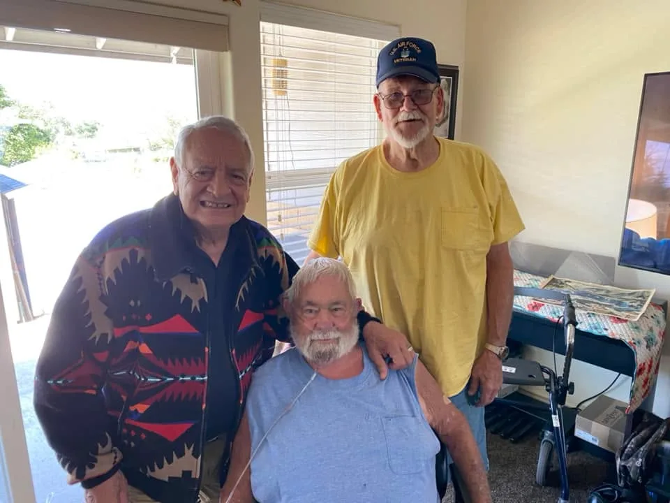 Phil Lane Jr. and classmates Gary Gilcher and Tim McCoy pose indoors: a man in a Native-patterned jacket, a seated man with an oxygen tube, and a man in a U.S. Air Force Veteran cap.