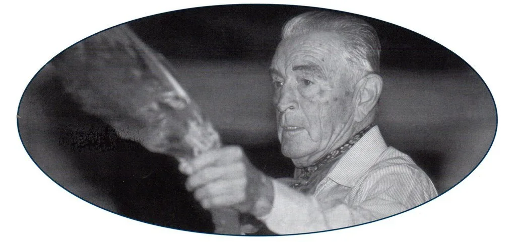 Black-and-white oval portrait of an elderly man with swept-back white hair and a patterned neckerchief, holding up a feathered object in motion.
