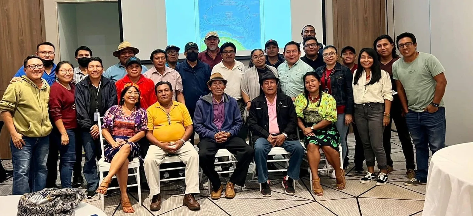 A large group of Guna men and women, some seated in front and others standing behind, pose together in a meeting room before a projected map.