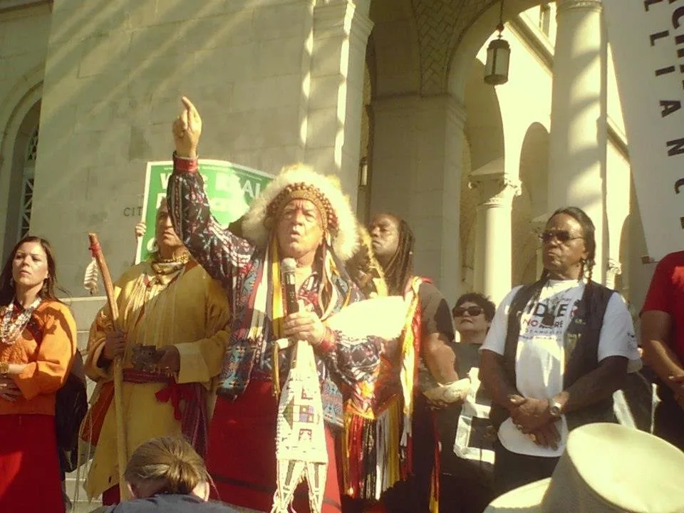 An elder in a large feathered headdress and patterned regalia raises one hand skyward while speaking into a microphone on the steps of a stone building, surrounded by others in traditional dress.