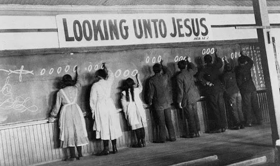 Historical black-and-white photograph of Indigenous children in uniforms facing a chalkboard, writing rows of zeros, beneath a banner reading "Looking Unto Jesus."