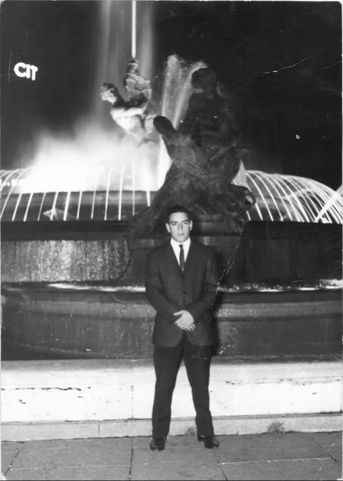 Black-and-white photo of a young man in a dark suit and tie standing before a lit fountain with bronze sculptures at night in Rome.