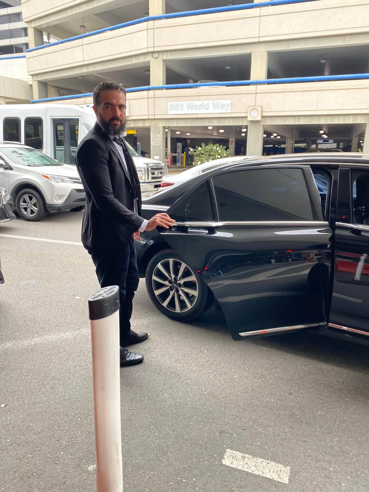 A bearded man in a black suit holds open the rear door of a black sedan at an airport curb, with a parking structure marked "551 World Way" behind him.