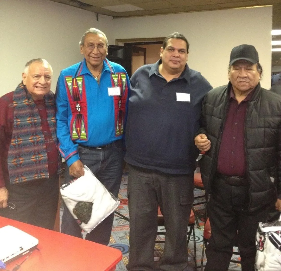 Four Indigenous men stand together in a meeting room, one in a blue ribbon shirt holding a white bundle, another in a patterned vest, one in a black cap.