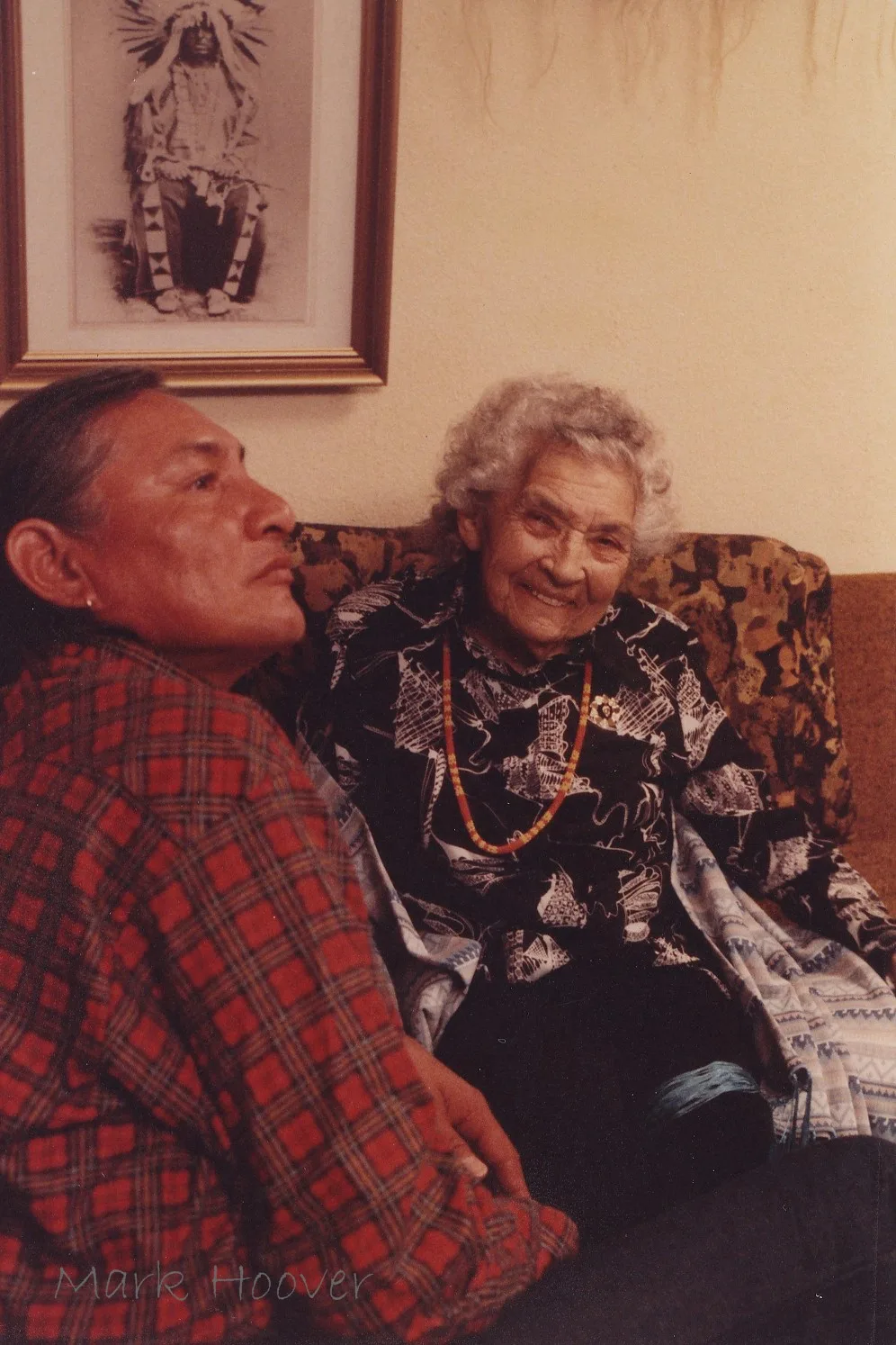 Will Sampson, in a red plaid shirt, sits beside a smiling white-haired Unchi Martina Sherwood wearing a beaded necklace; a framed portrait of a chief in a headdress hangs behind them.