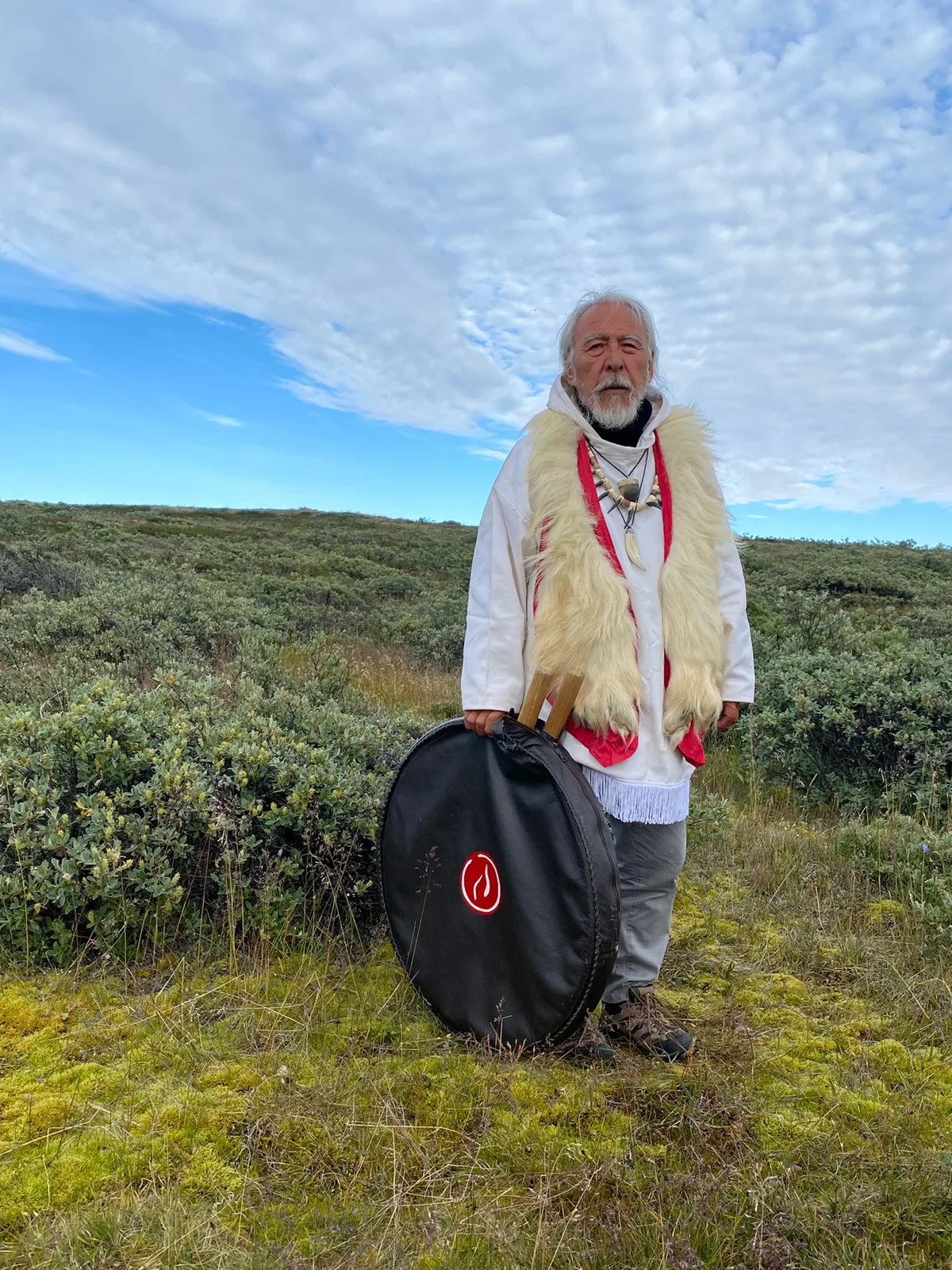 An elder with long white hair and a beard stands on green tundra under a cloudy blue sky, wearing a white tunic with a fur stole and bone necklace, holding a large drum in a black case.