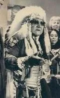 Black-and-white photograph of an elder in a large feather headdress, dark glasses and beaded regalia, speaking among a group of people.