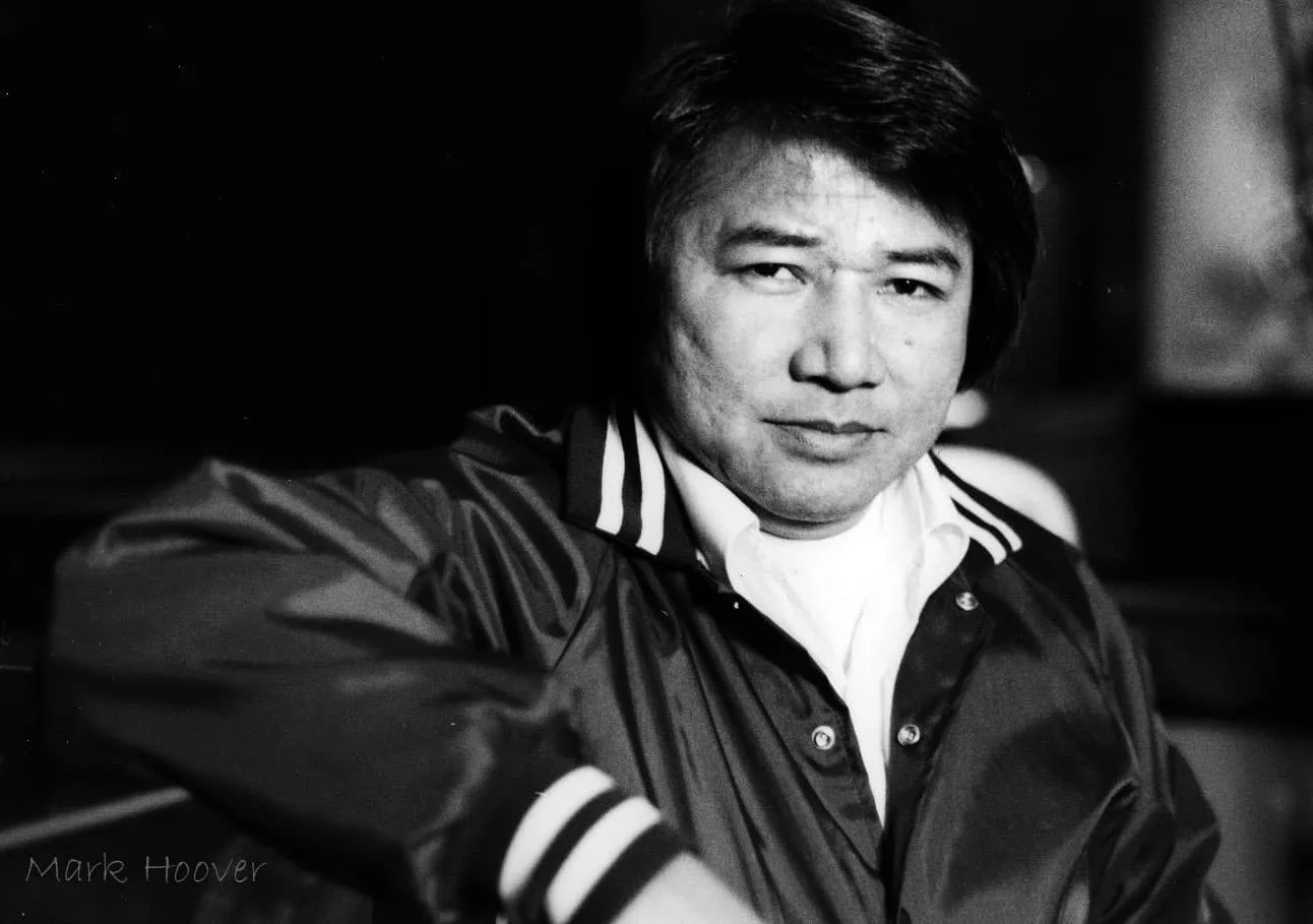 Black-and-white portrait of a man with dark hair in a satin jacket with striped collar and cuffs, gazing at the camera; photographer credit Mark Hoover in the corner.
