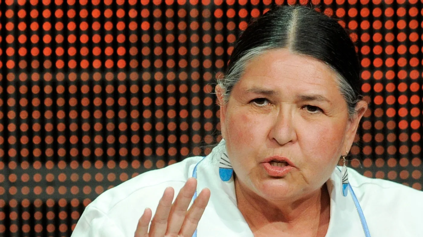 Sacheen, an older woman with gray-streaked dark hair pulled back, speaks and gestures with one hand, wearing a white top and blue-and-white earrings before an orange dotted backdrop.