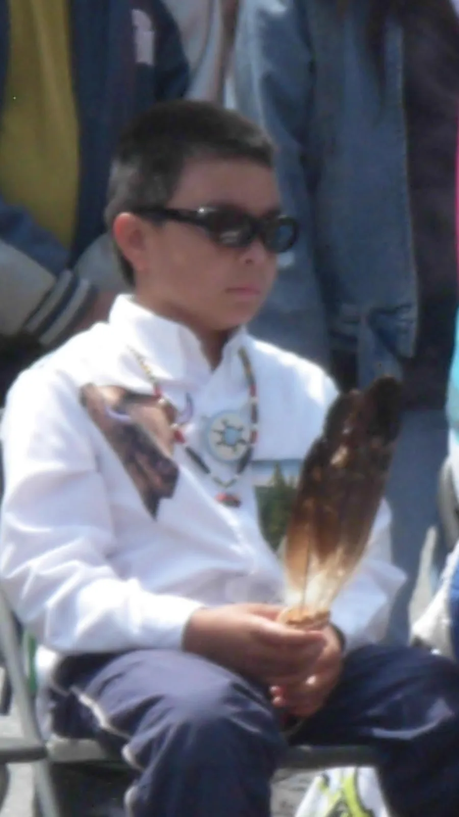 A young boy in dark sunglasses and a white shirt with beadwork and a medallion necklace sits holding a fan of eagle feathers, with people standing behind him.