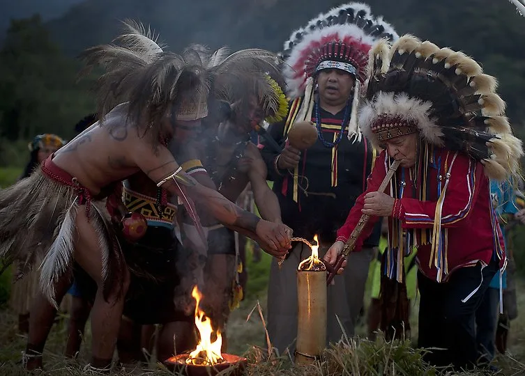 At dusk, Indigenous people in feathered headdresses and ceremonial dress gather around a small fire and a lit vessel, two elders in large eagle-feather war bonnets among them.