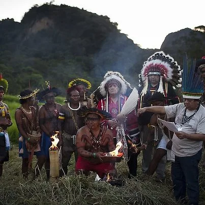 Indigenous leaders in feathered headdresses and regalia gather in a grassy field before forested hills at dusk, one kneeling with burning torches while another reads from papers.