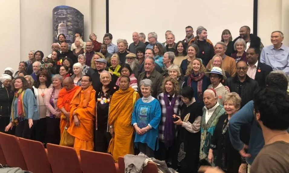 A large interfaith group poses together in an auditorium, including Buddhist monks in orange and saffron robes, elders and people of many backgrounds, with red seats in front.