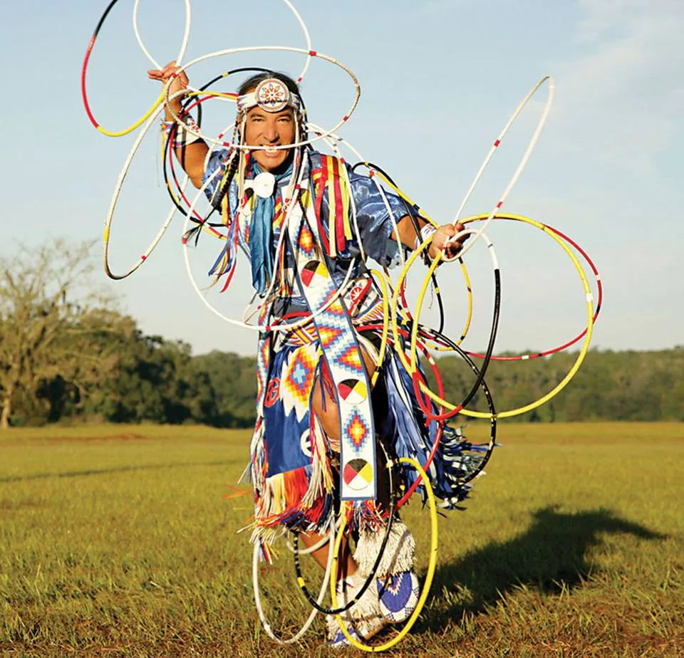 A smiling hoop dancer in blue regalia with beaded medicine wheel designs and fringe, spinning many red, yellow, white and black hoops in a sunlit grassy field.