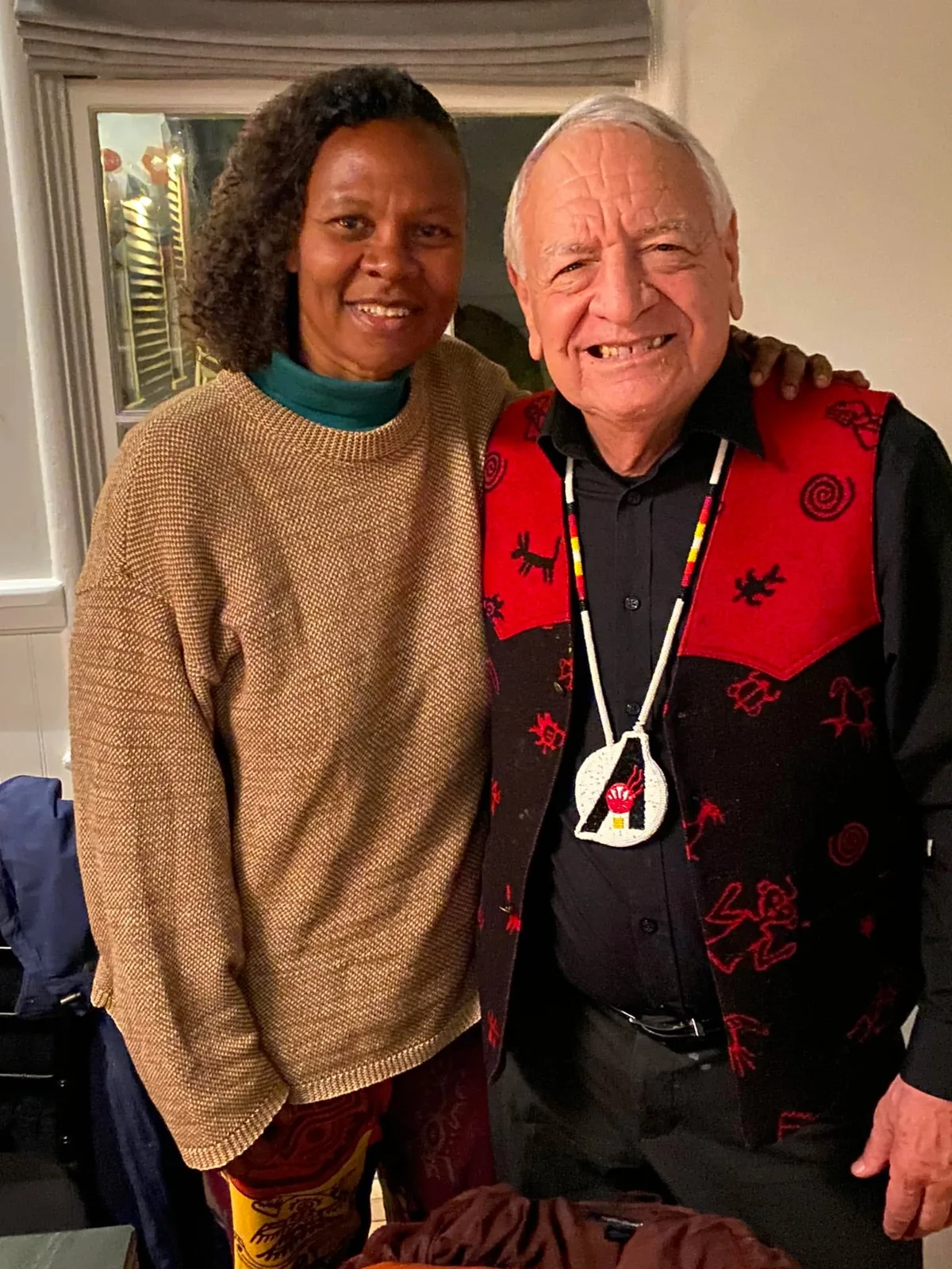 Dr. Yvonne Cagle, in a tan knit sweater, stands smiling with her arm around Phil Lane Jr., who wears a red and black vest with Indigenous symbols and a beaded medallion.