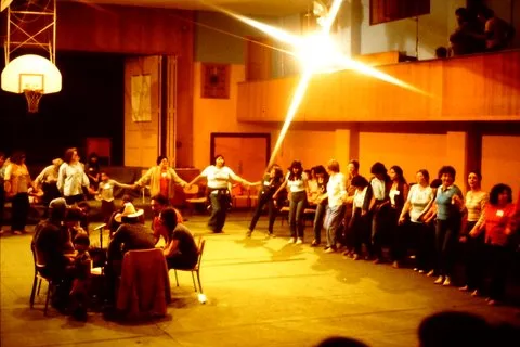 A large group of people holding hands in a long line, dancing in a circle around a gymnasium hall with a basketball hoop, while others sit at a table nearby.