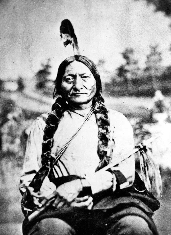 Historic black-and-white portrait of Sitting Bull, seated, with a single feather in his hair, fur-wrapped braids, a beaded strap across his chest, holding a pipe.