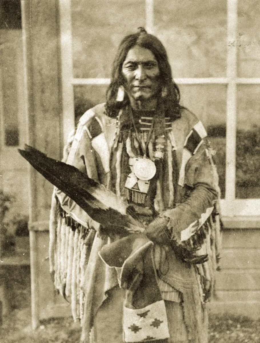 Sepia historical portrait of Chief Crowfoot standing before a window, wearing a fringed hide shirt and a large medallion, holding an eagle feather fan and a beaded bag.