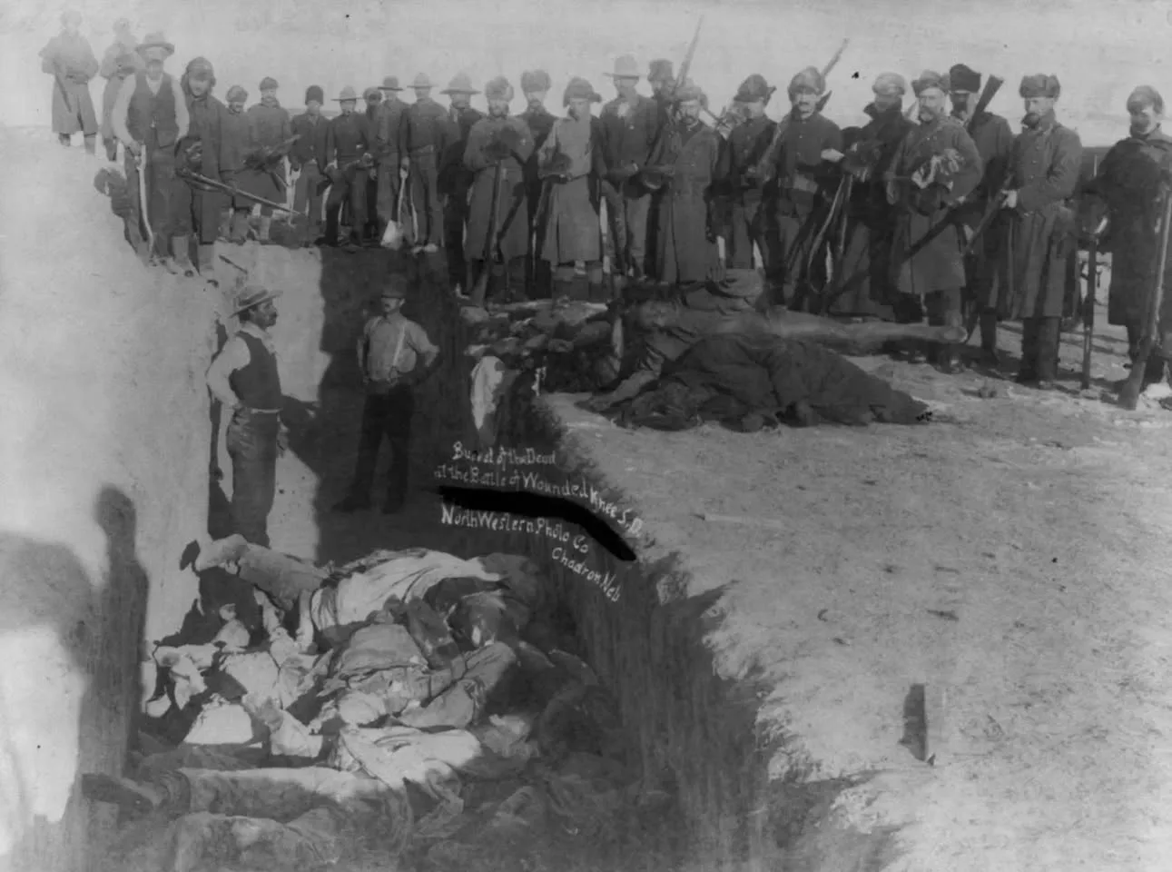 Historic black-and-white photograph of soldiers and civilians with rifles and shovels standing beside a mass grave of Lakota dead at Wounded Knee in winter, 1890.