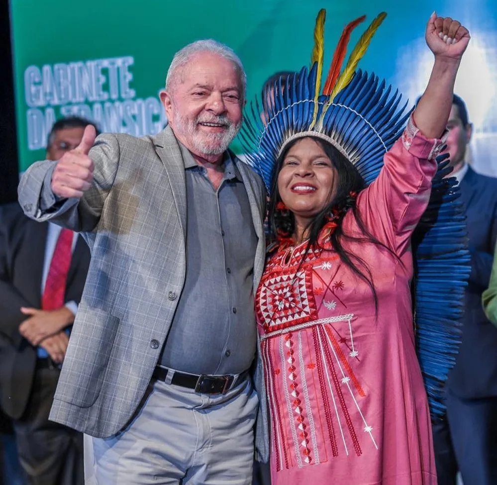 President-elect Lula, smiling and giving a thumbs up, stands beside Sônia Guajajara, who raises her fist, wearing a blue feather headdress and an embroidered pink dress.
