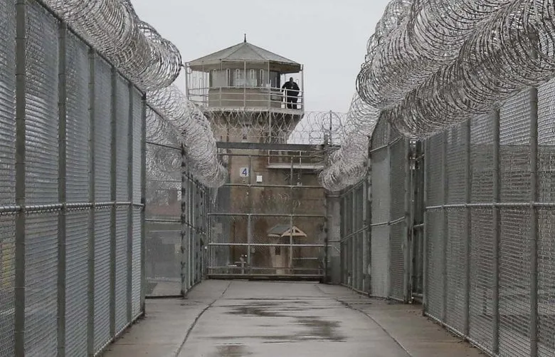 A walkway between tall chain-link fences topped with coiled razor wire leads to a prison guard tower, where a guard stands on the upper balcony.