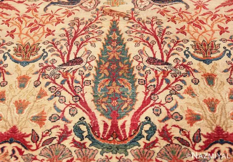 Close-up of an antique Persian carpet with a green cypress tree at its center, framed by red flowering branches, perched birds and lotus blossoms on an ivory field.