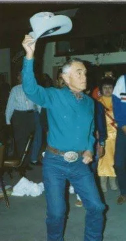A gray-haired man in a teal shirt, jeans and a large belt buckle raises a light-colored cowboy hat high while dancing at an indoor gathering, with people standing behind him.