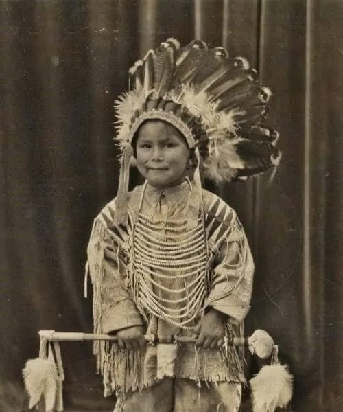 Sepia studio portrait of young Emil J. Comes Last in a feathered war bonnet and fringed buckskin shirt with a bone breastplate, holding a decorated pipe.