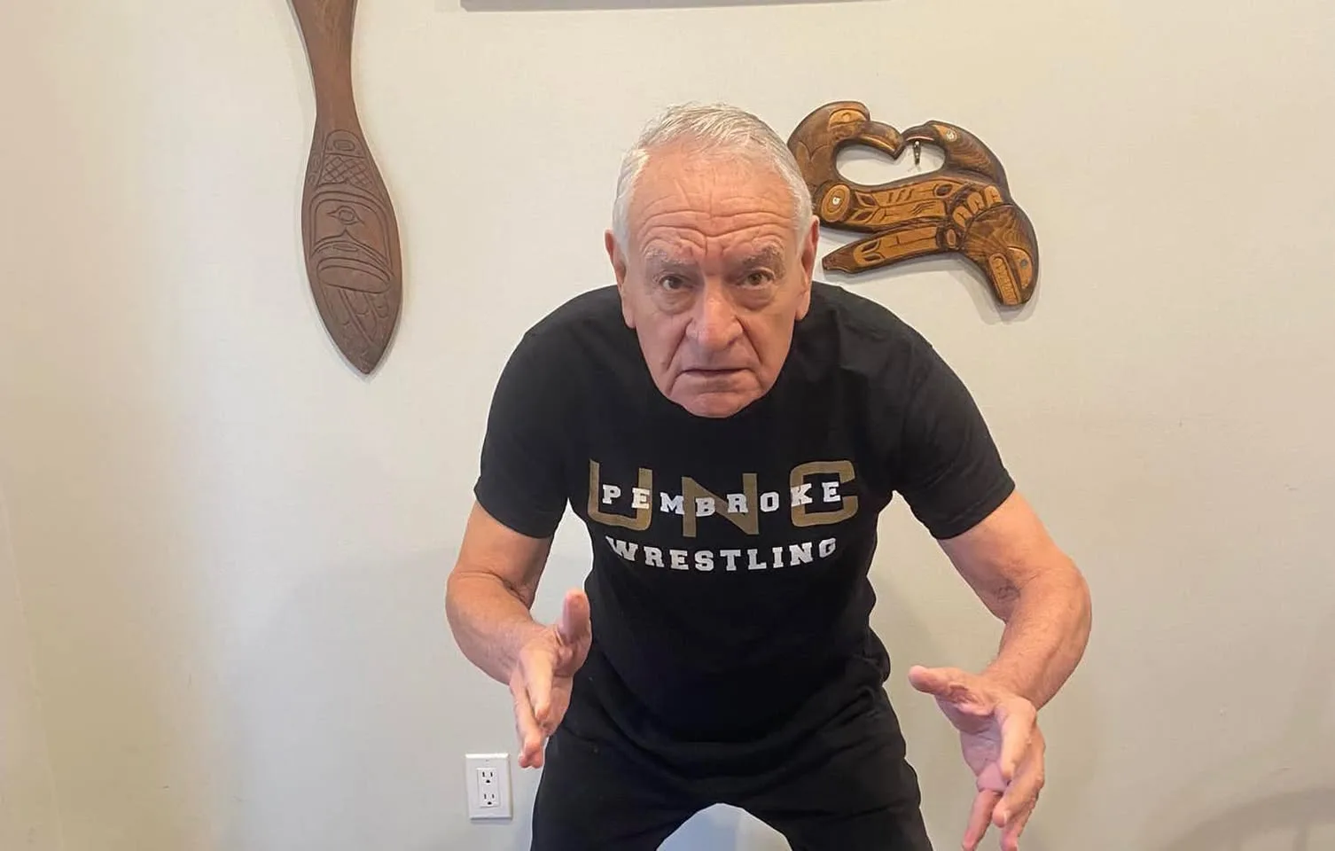 A white-haired elder in a black UNC Pembroke Wrestling T-shirt crouches in a wrestling stance, hands ready, before a wall with carved Northwest Coast paddle and bird art.