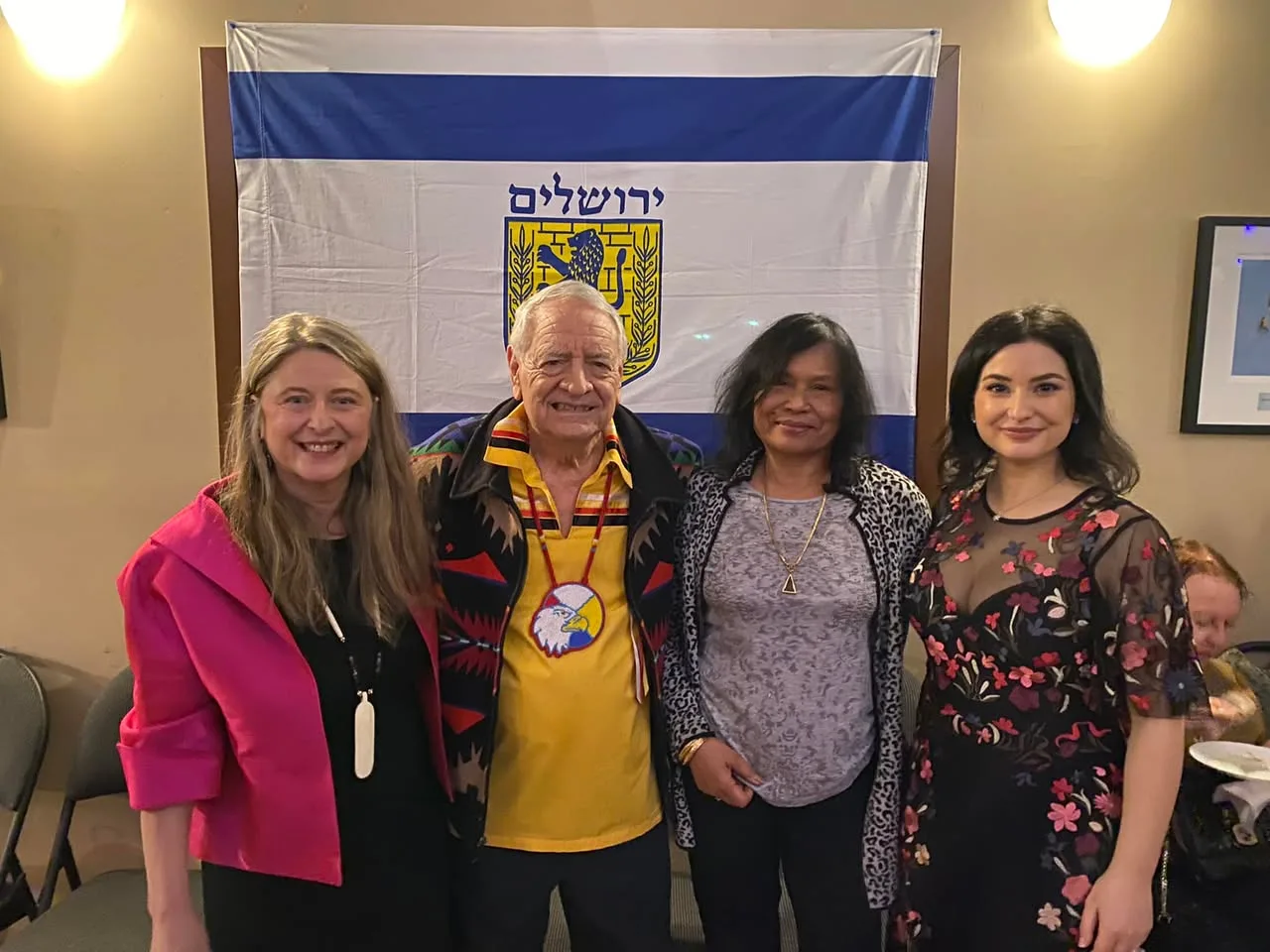 Phil Lane Jr., in a yellow ribbon shirt and eagle medallion, stands with three women before a Jerusalem flag at the White Rock-Surrey Jewish Community Centre.