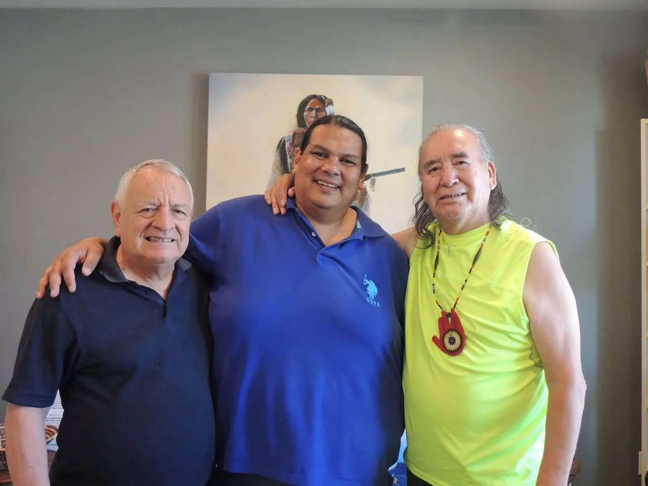 Three smiling men stand arm in arm in front of a painting of a Native warrior holding a rifle; the man on the right wears a beaded red hand medallion.