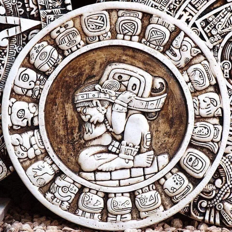 A carved circular stone disc in Mayan style showing a seated figure in an elaborate headdress at the center, surrounded by a ring of calendar glyphs.