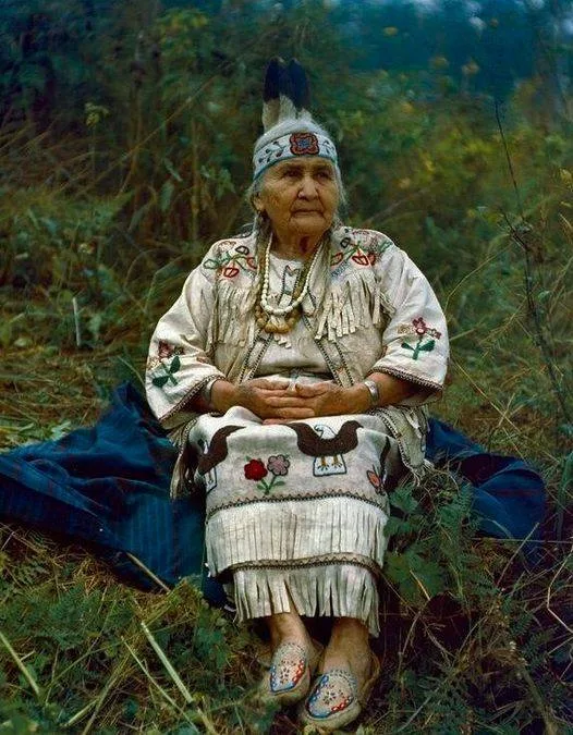 Mrs. Taha George, an elder, seated on a blanket in tall grass, wearing a fringed white buckskin dress with floral and bird beadwork, a beaded headband with feathers and beaded moccasins.