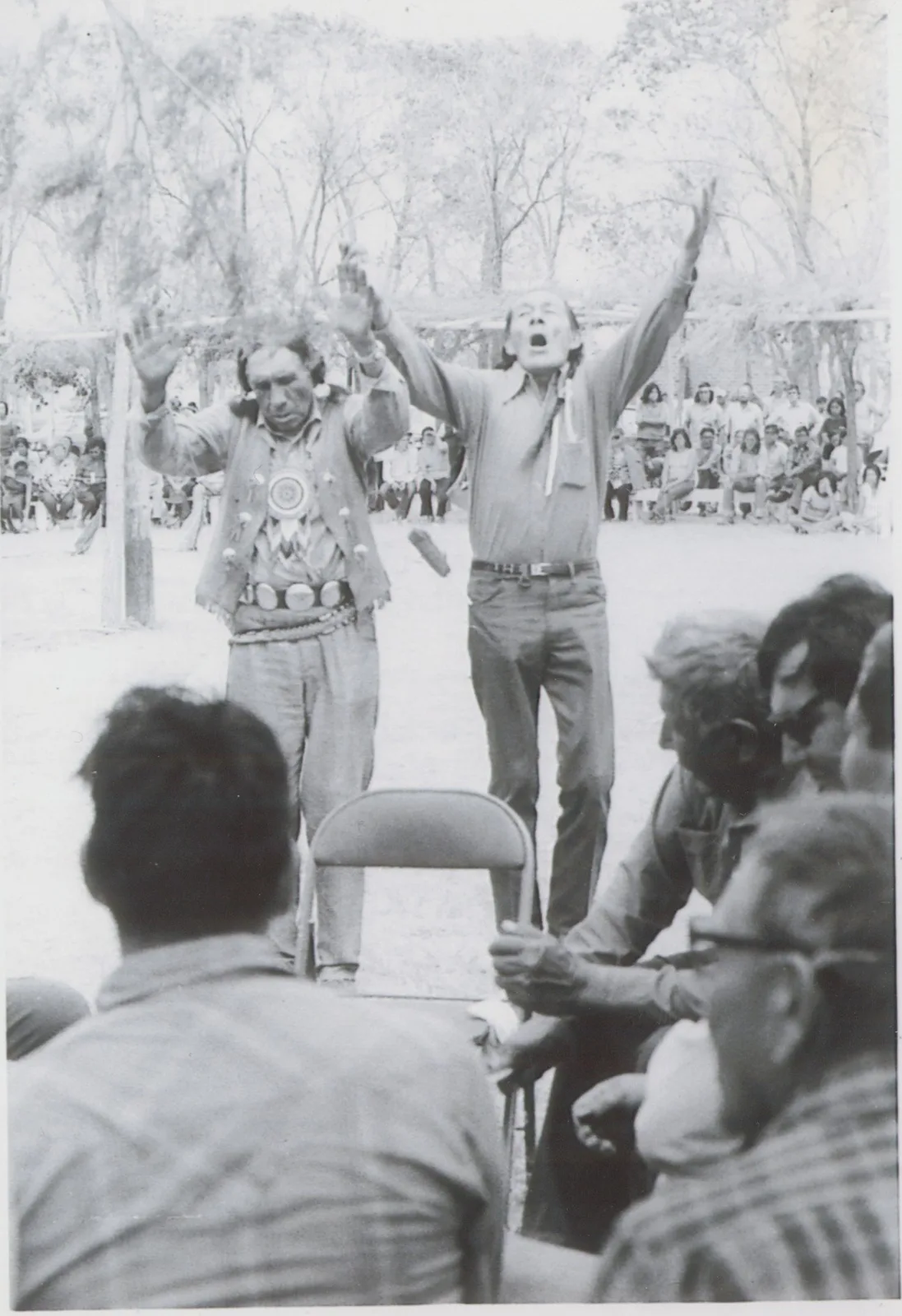 Black-and-white photograph of two elders standing with arms raised before seated people at an outdoor gathering. On the left, John Fire Lame Deer in a beaded vest and concho belt; on the right, Pete Catches Sr.