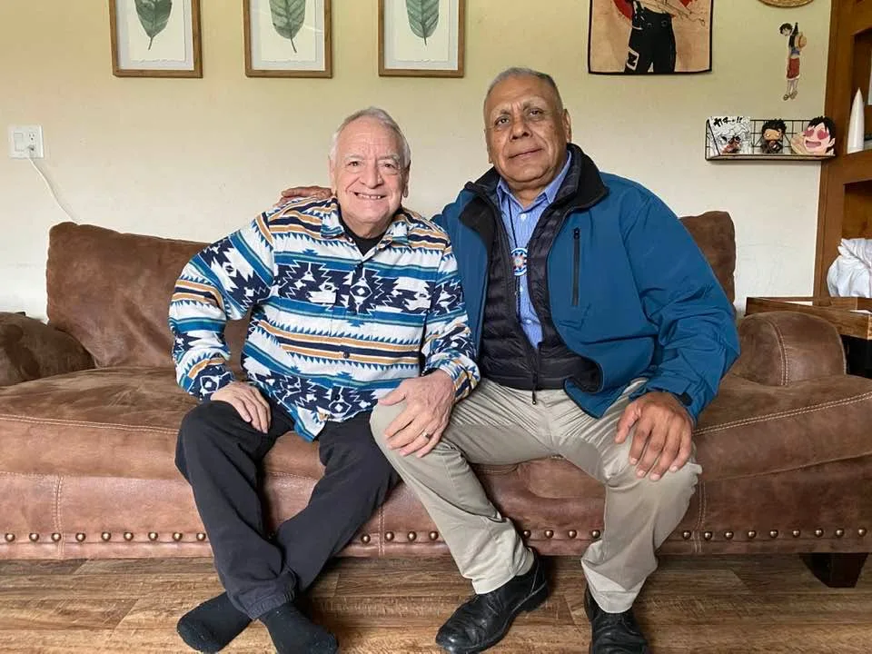 Two men sit side by side on a brown leather couch, smiling, one in a blue and white patterned shirt and the other, his arm around him, in a blue jacket.